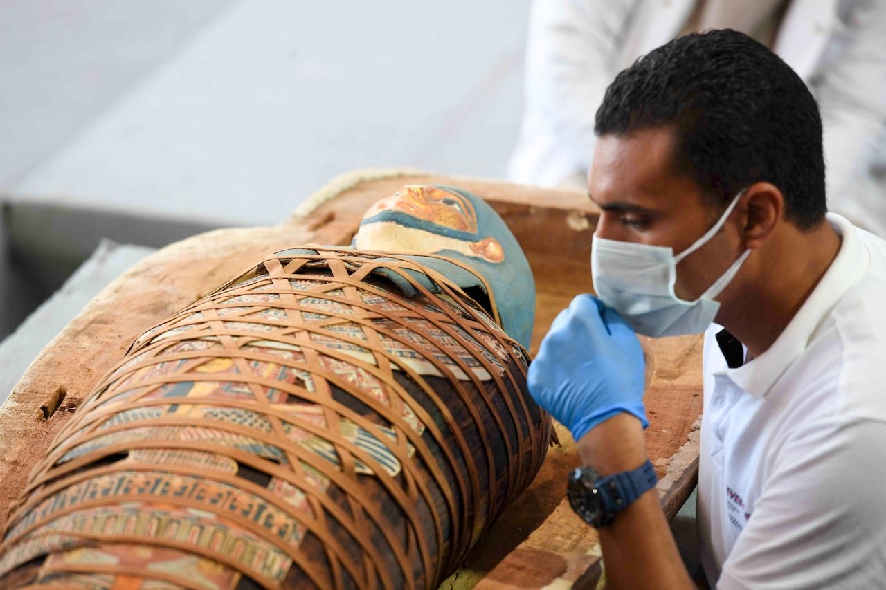 default-783 An archaeologist inspects an Egyptian mummy that is wrapped in an elaborate and colourful burial shroud. The archaeologist is wearing a medical mask and gloves.