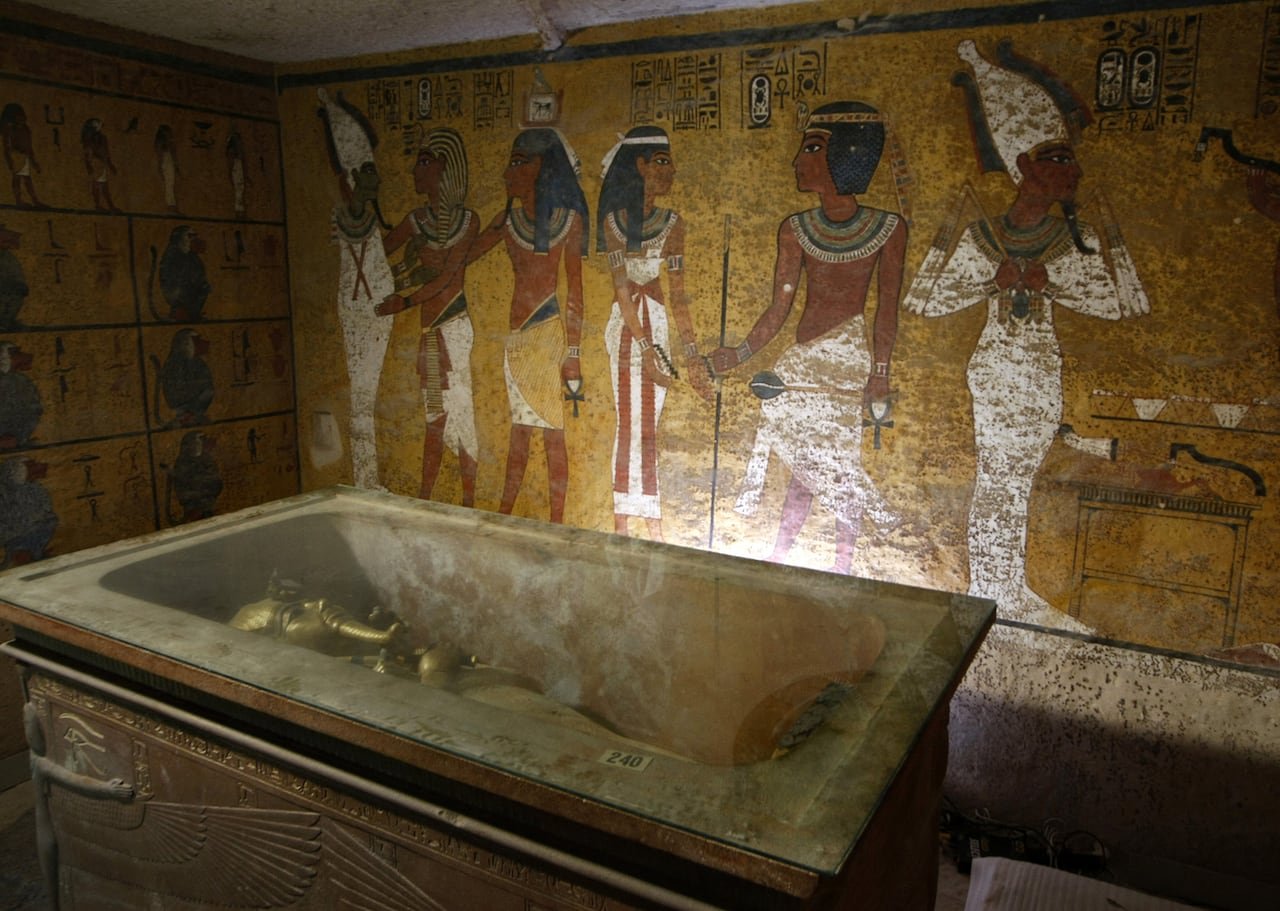 default-784 A golden sarcophagus is shown lying in an elaborate burial chamber. The wall behind it is colourfully painted with figures.