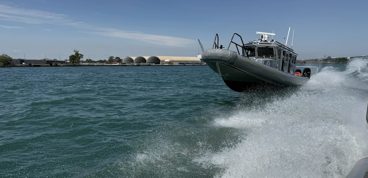 default-26 A grey-roofed boat is depicted navigating waves in a river, with sea spray emanating from another passing vessel.